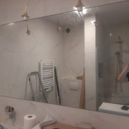 Michał Cader - Duże lustro w łazience z białymi marmurkowymi płytkami, odbijające grzejnik, drabinę i oświetlenie, z widocznym fotografem robiącym zdjęcie w odbiciu.