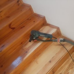 Michał Cader - Drewniane schody w trakcie cyklinowania, widoczna szlifierka oscylacyjna z odkurzaczem, fragment schodów po obróbce jaśniejszy, ukazujący odnowioną powierzchnię drewna.