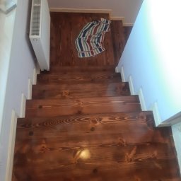 Michał Cader - Wykończone drewniane schody z widocznymi słojami, białe listwy przypodłogowe, grzejnik na ścianie i ręcznik w paski na podłodze na górze schodów.