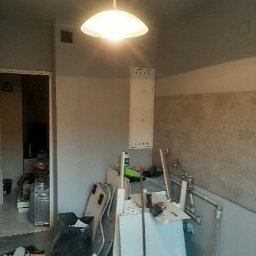 Michał Cader - Kuchnia w trakcie remontu: widoczne niedokończone ściany, porozrzucane materiały budowlane, biała lampa sufitowa i fragmenty mebli.