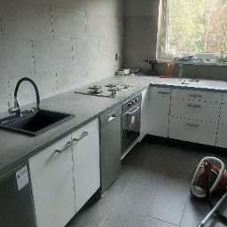 Michał Cader - Nowoczesna kuchnia po remoncie: szare płytki na ścianie, czarny zlew z baterią, białe szafki z szarym blatem, płyta grzewcza, piekarnik i zmywarka. Na blacie przy oknie widać wiertarkę i akcesoria...