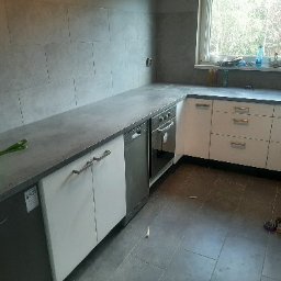 Michał Cader - Wykończona kuchnia z szarymi płytkami na ścianie i podłodze, białe szafki z poziomymi uchwytami, widoczne narzędzia i akcesoria na blacie i podłodze.