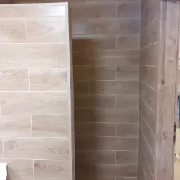 Ukończona łazienka z kabiną prysznicową wyłożona płytkami imitującymi drewno, widoczna kratka wentylacyjna i fragment ościeżnicy drzwi.