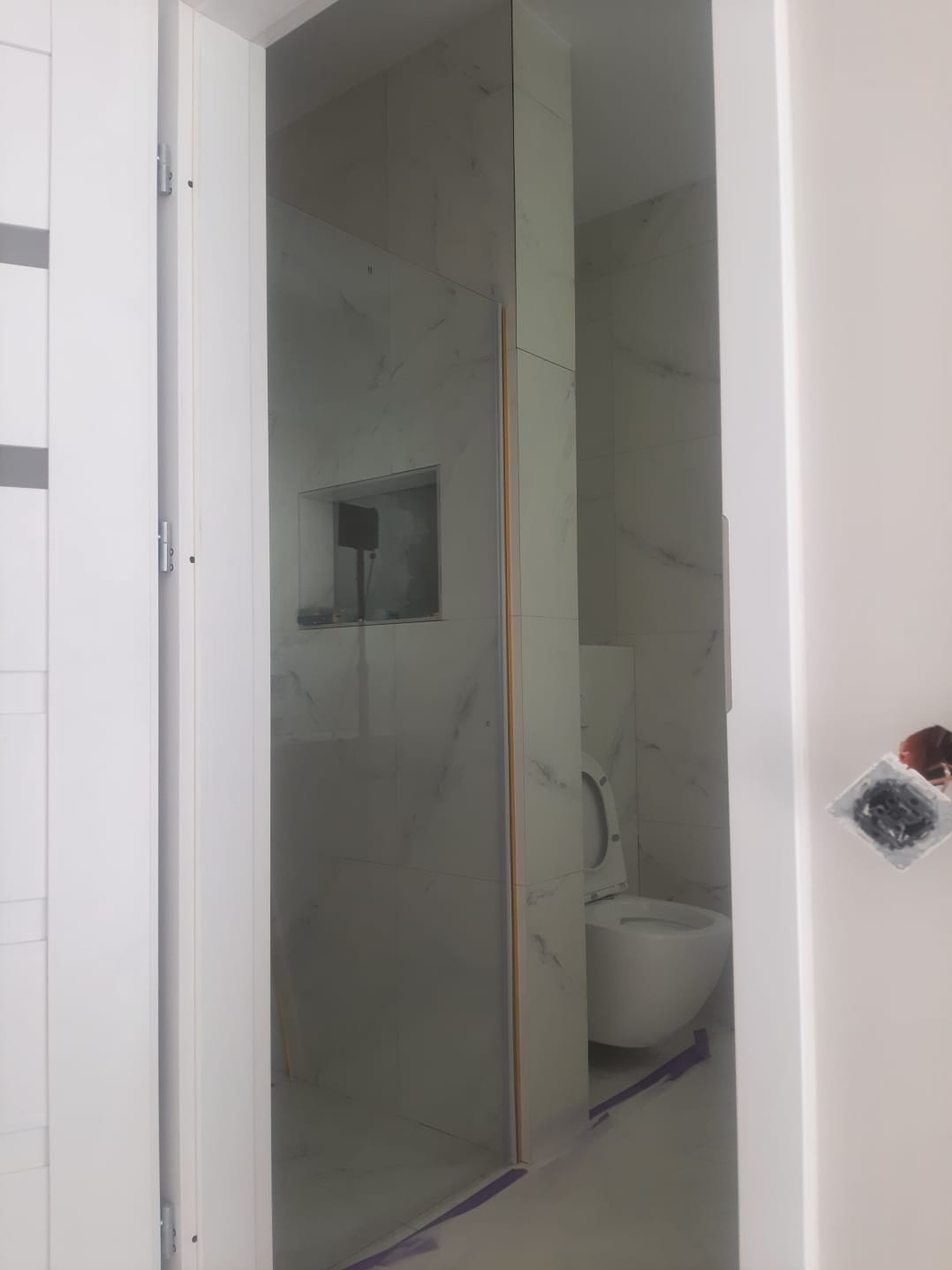 Wnętrze łazienki w trakcie wykańczania: białe marmurowe płytki, kabina prysznicowa z widocznym profilem, toaleta w stylu nowoczesnym, zabezpieczenia podłogi folią malarską, gniazdko elektryczne bez...