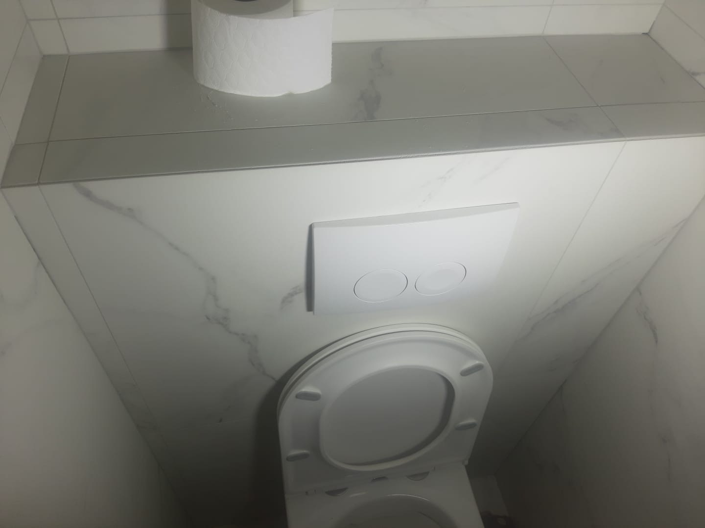 Toaleta wisząca z białą deską i przyciskiem spłukującym na ścianie wyłożonej białymi płytkami imitującymi marmur, nad którą znajduje się rolka papieru toaletowego.