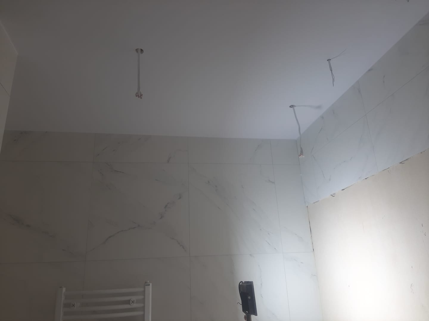 Remont łazienki w trakcie: położone płytki imitujące marmur na ścianach, widoczne kable oświetleniowe wystające z sufitu, fragment ściany przed wykończeniem.