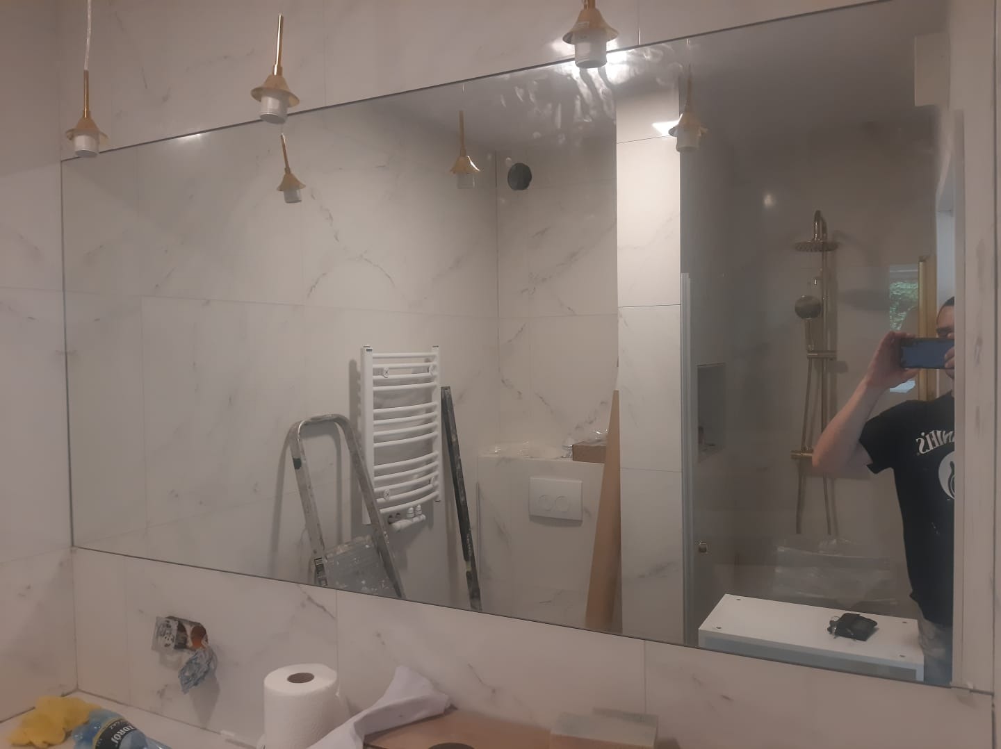 Duże lustro w łazience z białymi marmurkowymi płytkami, odbijające grzejnik, drabinę i oświetlenie, z widocznym fotografem robiącym zdjęcie w odbiciu.