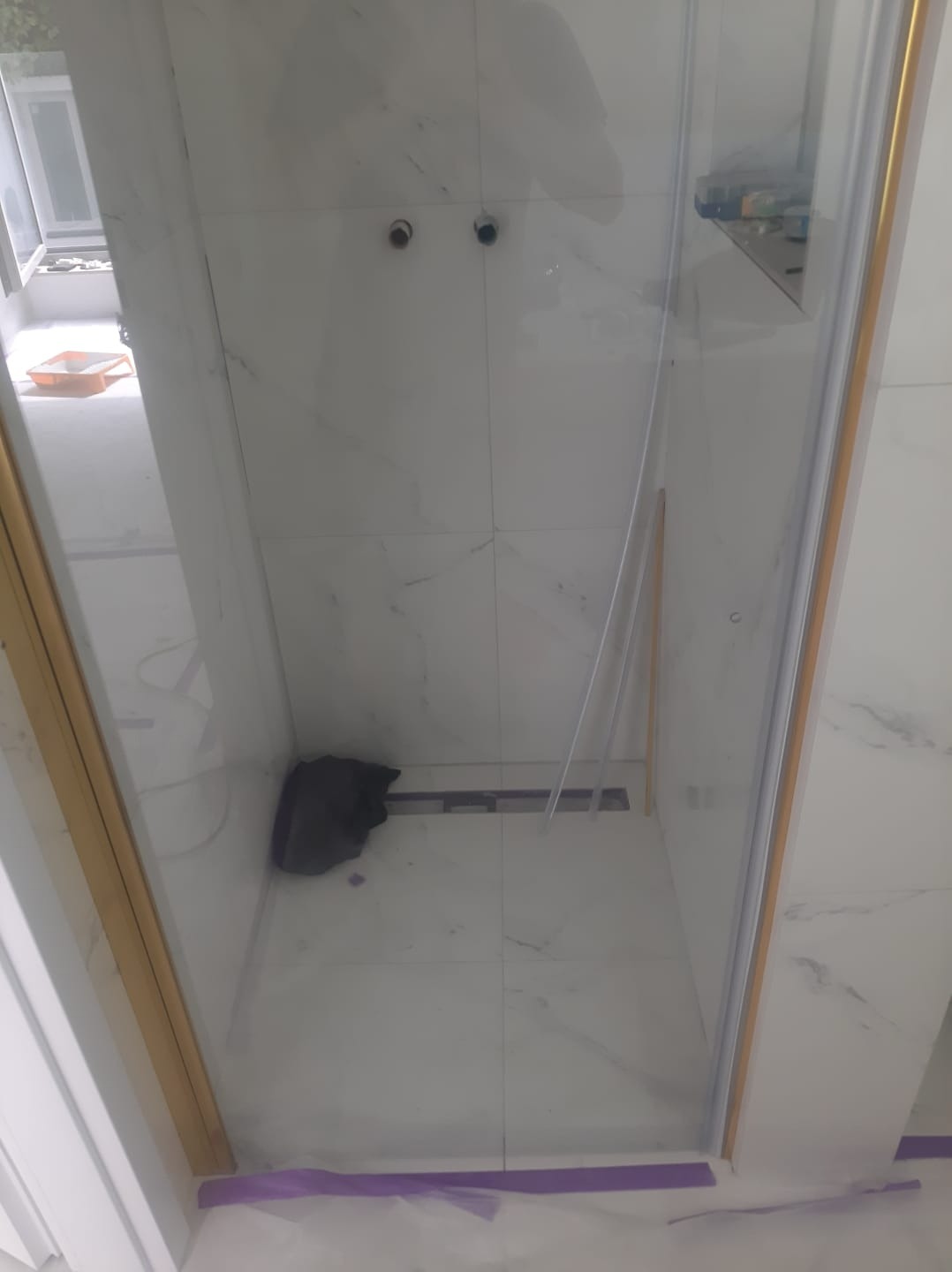 Kabina prysznicowa z złotymi ramami i odpływem liniowym, zabezpieczona folią malarską u dołu, w trakcie prac wykończeniowych w łazience z białymi marmurkowymi płytkami.