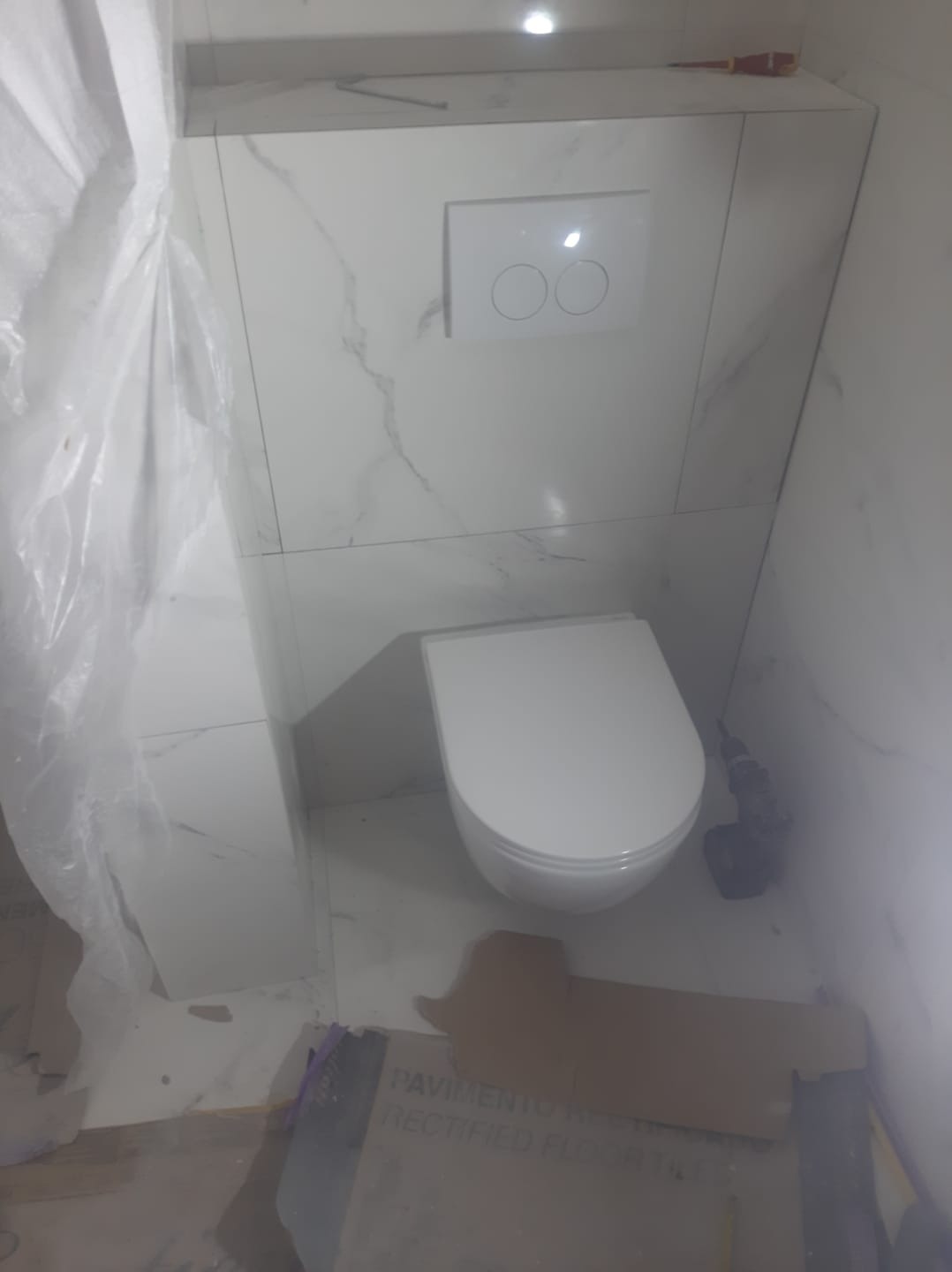 Wisząca toaleta z podtynkową spłuczką na tle marmurowych płytek, obok wiertarka i folia ochronna, w trakcie prac wykończeniowych.