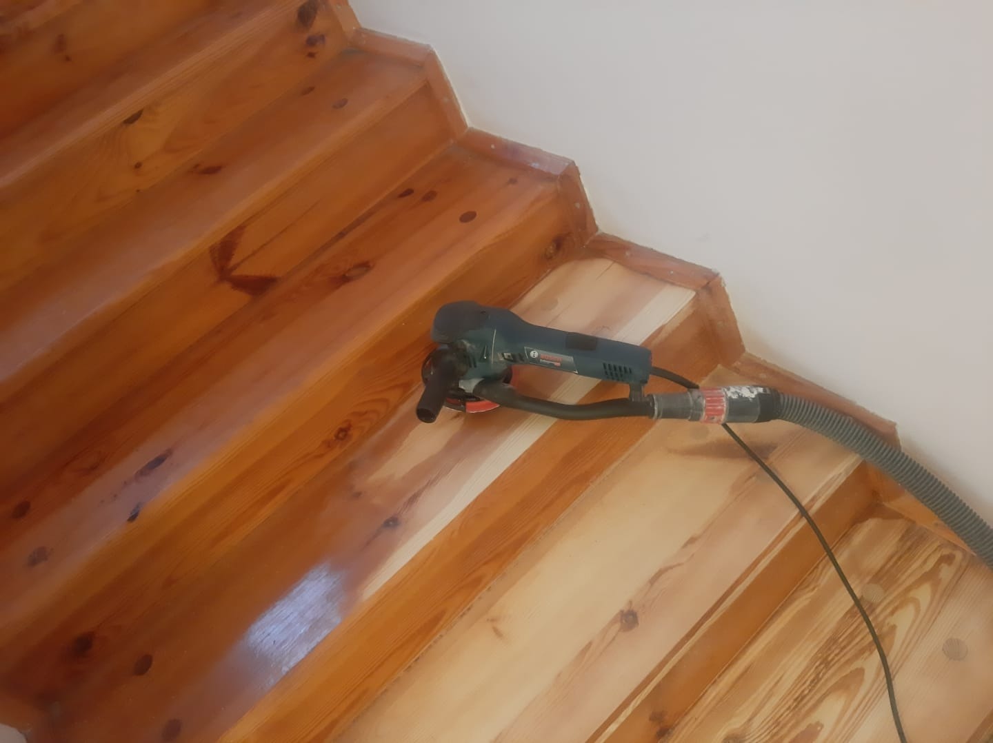 Drewniane schody w trakcie cyklinowania, widoczna szlifierka oscylacyjna z odkurzaczem, fragment schodów po obróbce jaśniejszy, ukazujący odnowioną powierzchnię drewna.