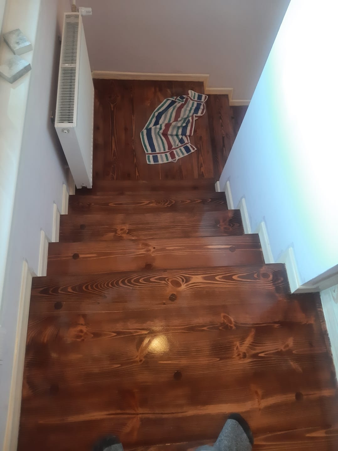 Wykończone drewniane schody z widocznymi słojami, białe listwy przypodłogowe, grzejnik na ścianie i ręcznik w paski na podłodze na górze schodów.