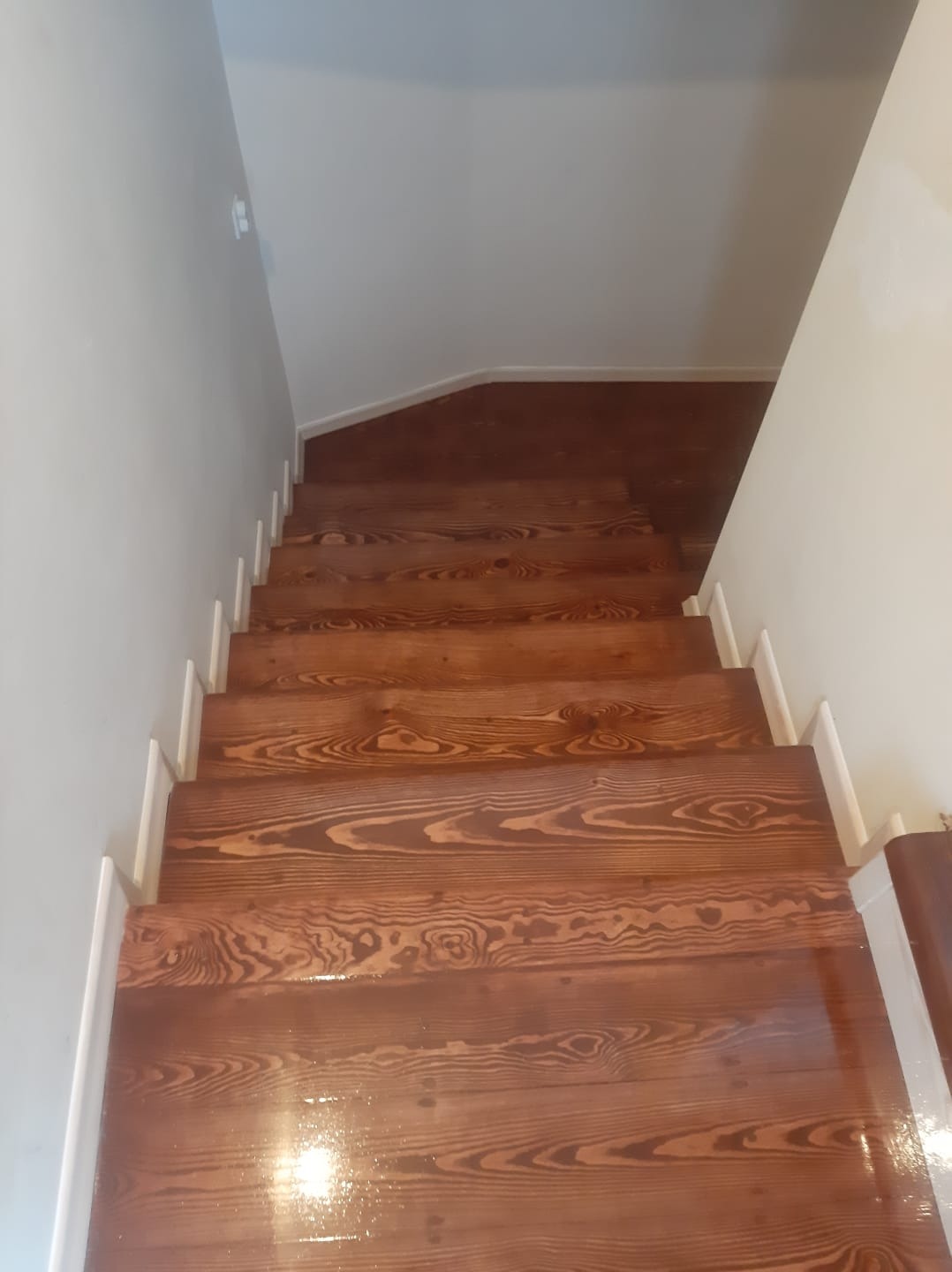 Odnowione drewniane schody z widocznym usłojeniem, pomalowane na brązowo, z białymi listwami przypodłogowymi i kremowymi ścianami, sfotografowane z góry.