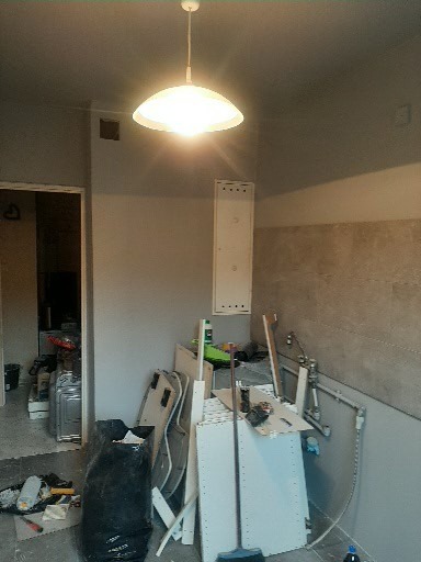 Kuchnia w trakcie remontu: widoczne niedokończone ściany, porozrzucane materiały budowlane, biała lampa sufitowa i fragmenty mebli.