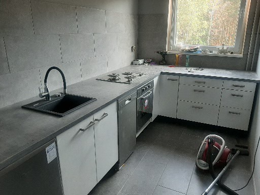 Nowoczesna kuchnia po remoncie: szare płytki na ścianie, czarny zlew z baterią, białe szafki z szarym blatem, płyta grzewcza, piekarnik i zmywarka. Na blacie przy oknie widać wiertarkę i akcesoria...