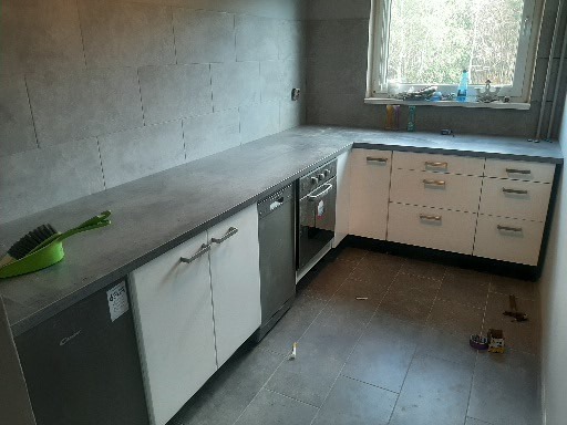 Wykończona kuchnia z szarymi płytkami na ścianie i podłodze, białe szafki z poziomymi uchwytami, widoczne narzędzia i akcesoria na blacie i podłodze.