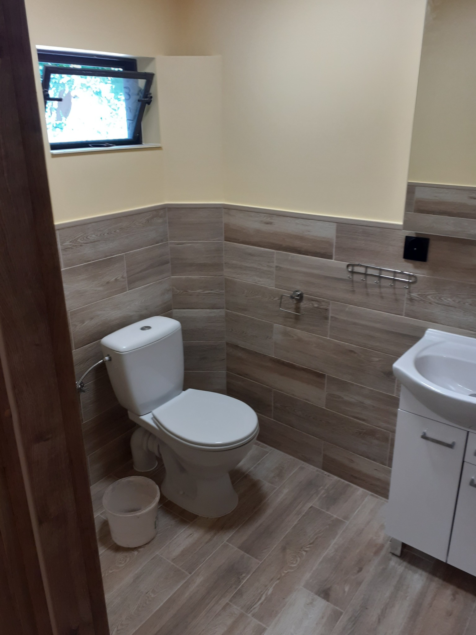 Jasna łazienka po remoncie z drewnopodobnymi płytkami, białą armaturą i uchylnym oknem piwnicznym.