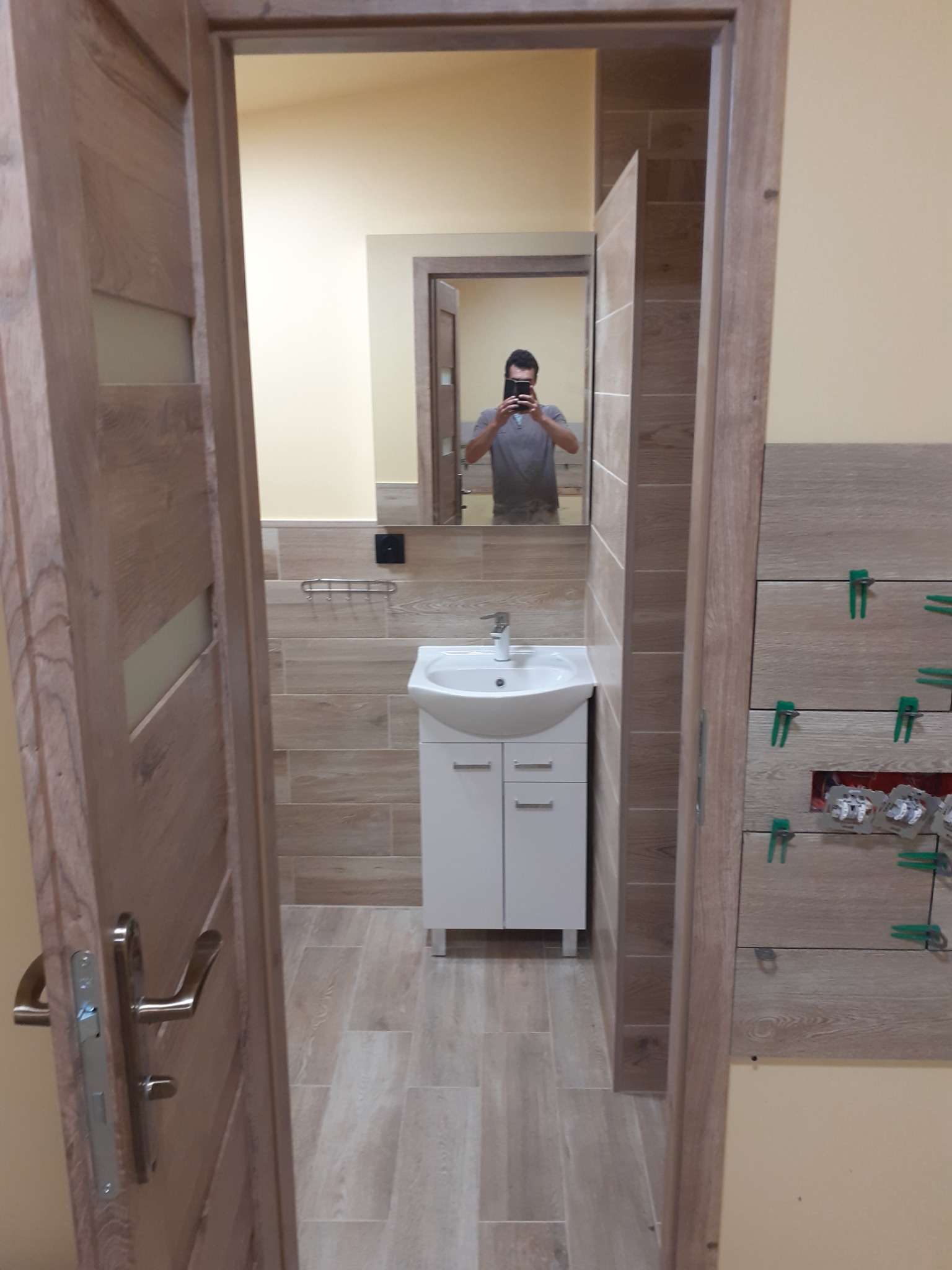 Łazienka w trakcie wykańczania: widoczne płytki imitujące drewno na ścianach, biała szafka pod umywalką, lustro z odbiciem fotografa i zielone krzyżyki dystansowe między płytkami na ścianie po...