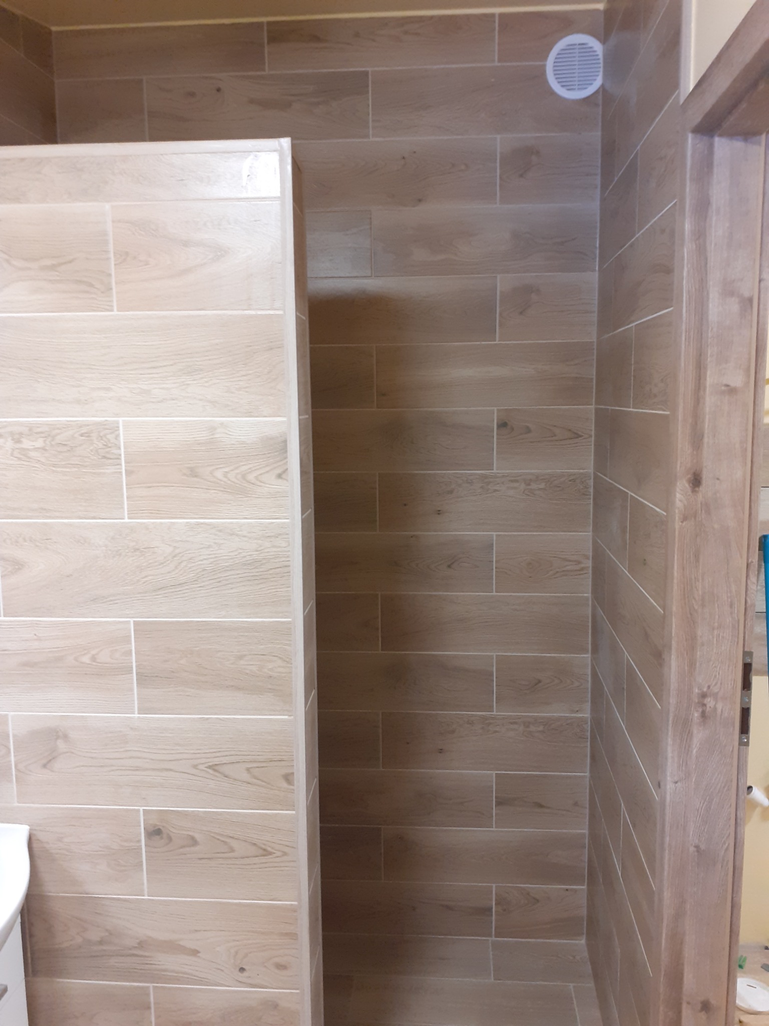 Ukończona łazienka z kabiną prysznicową wyłożona płytkami imitującymi drewno, widoczna kratka wentylacyjna i fragment ościeżnicy drzwi.