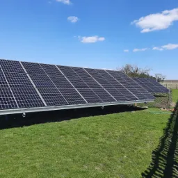 Panele fotowoltaiczne zamontowane na trawniku, widoczne mocowania i cień rzucany przez ogrodzenie, błękitne niebo z chmurami w tle.