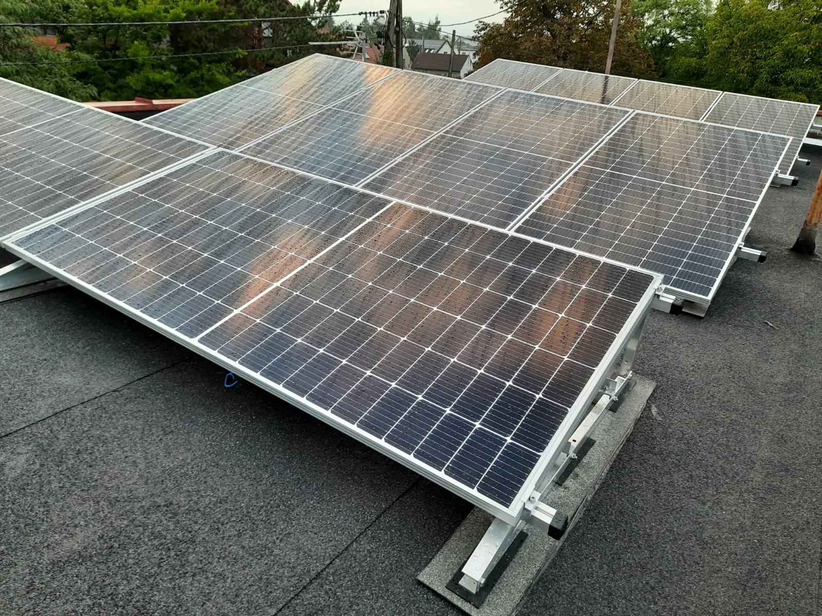 Panel słoneczny z kroplami deszczu na czarnym dachu, zamontowany na aluminiowej konstrukcji z szarymi podkładkami antywibracyjnymi, widok z góry.
