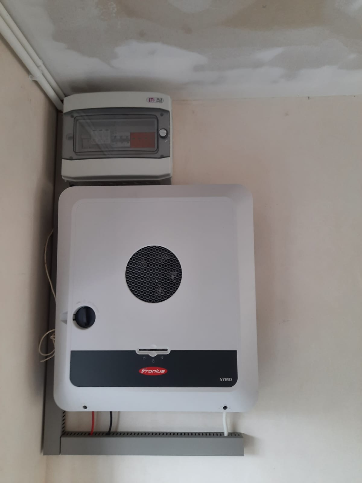 Biały falownik Fronius SYMO zamontowany na ścianie, widoczna skrzynka elektryczna powyżej, instalacja w surowym stanie.