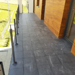 Szare płytki gresowe na balkonie z drewnopodobną elewacją i metalowymi barierkami, widok na zielone pola w oddali.