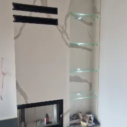 Nowoczesny kominek z marmurowym wykończeniem, czarnymi wstawkami i szklanymi półkami po prawej stronie, surowy kabel zwisający z sufitu, podłoga z paneli.