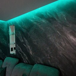BP Błażej Pietrzak - Ściana z dekoracyjną fakturą imitującą marmur, podświetlona zielonym światłem LED, nad szarą sofą z poduszkami. W tle widoczna szklana półka z dekoracjami.