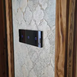 Dekoracyjna ściana z wzorem marokańskim w odcieniach szarości i beżu, z nowoczesnym dotykowym włącznikiem światła z niebieskimi diodami, obok drewniana futryna drzwi.