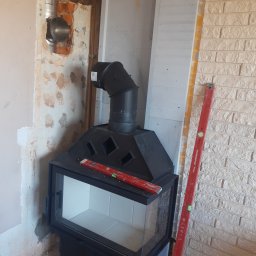 BP Błażej Pietrzak - Czarne żeliwne wkłady kominkowe z szybą, etap montażu z widocznym podłączeniem do komina, poziomica na górze, surowe ściany i elementy wykończeniowe w tle.