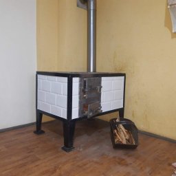 BP Błażej Pietrzak - Tradycyjny piec kaflowy z otwartymi drzwiczkami, metalowym kominem i koszem drewna obok, umieszczony w rogu pomieszczenia z drewnianą podłogą i ścianami w odcieniach bieli i żółci.