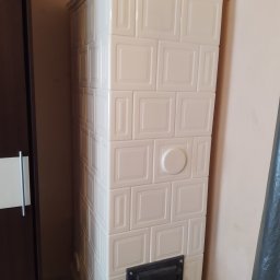 BP Błażej Pietrzak - Biały piec kaflowy z czarnymi drzwiczkami paleniska, widoczny obok ciemnych drzwi szafy, na tle jasnej ściany.