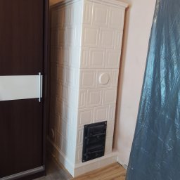 BP Błażej Pietrzak - Biały, kaflowy piec kominkowy z czarnymi drzwiczkami, ustawiony w rogu pokoju obok ciemnej szafy przesuwnej i przykryty folią malarską.