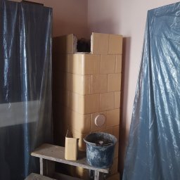 BP Błażej Pietrzak - W trakcie budowy pieca kaflowego w rogu pokoju, zabezpieczonego folią malarską, widoczny fragment rusztowania i wiadro z zaprawą.