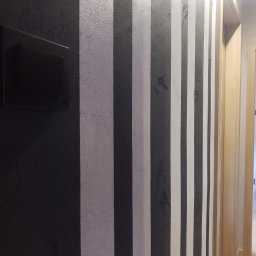 BP Błażej Pietrzak - Ściana z dekoracyjnym malowaniem w pionowe pasy w odcieniach szarości i bieli, obok włącznik światła i fragment drewnianej ościeżnicy drzwiowej.