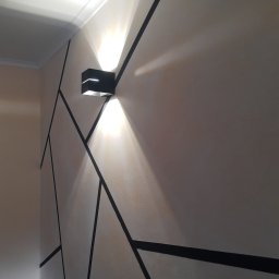 BP Błażej Pietrzak - Ściana z geometrycznym wzorem z czarnych linii i nowoczesnym kinkietem emitującym ciepłe światło.