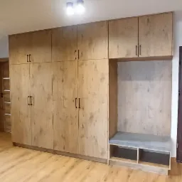 Duża szafa z jasnego drewna z ciemnymi uchwytami, wbudowana wnęka z siedziskiem obitym szarą tkaniną, podłoga z paneli drewnopodobnych.