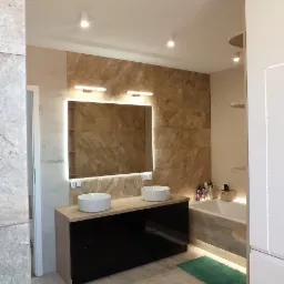 Nowoczesna łazienka z podwójną umywalką nablatową, szafką z fornirem drewnopodobnym i czarnymi frontami, podświetlanym lustrem i płytkami imitującymi kamień na ścianie.