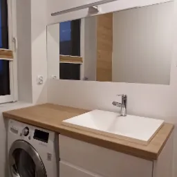 Biała umywalka nablatowa z drewnianym blatem, zamontowana na białej szafce z szufladami, obok pralka w łazience z lustrem i oświetleniem.