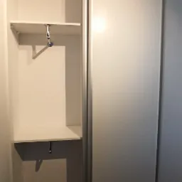 Wnętrze białej szafy wnękowej z przesuwnymi drzwiami, widoczne półki i drążek na ubrania, brązowa podłoga, gniazdko elektryczne na ścianie.