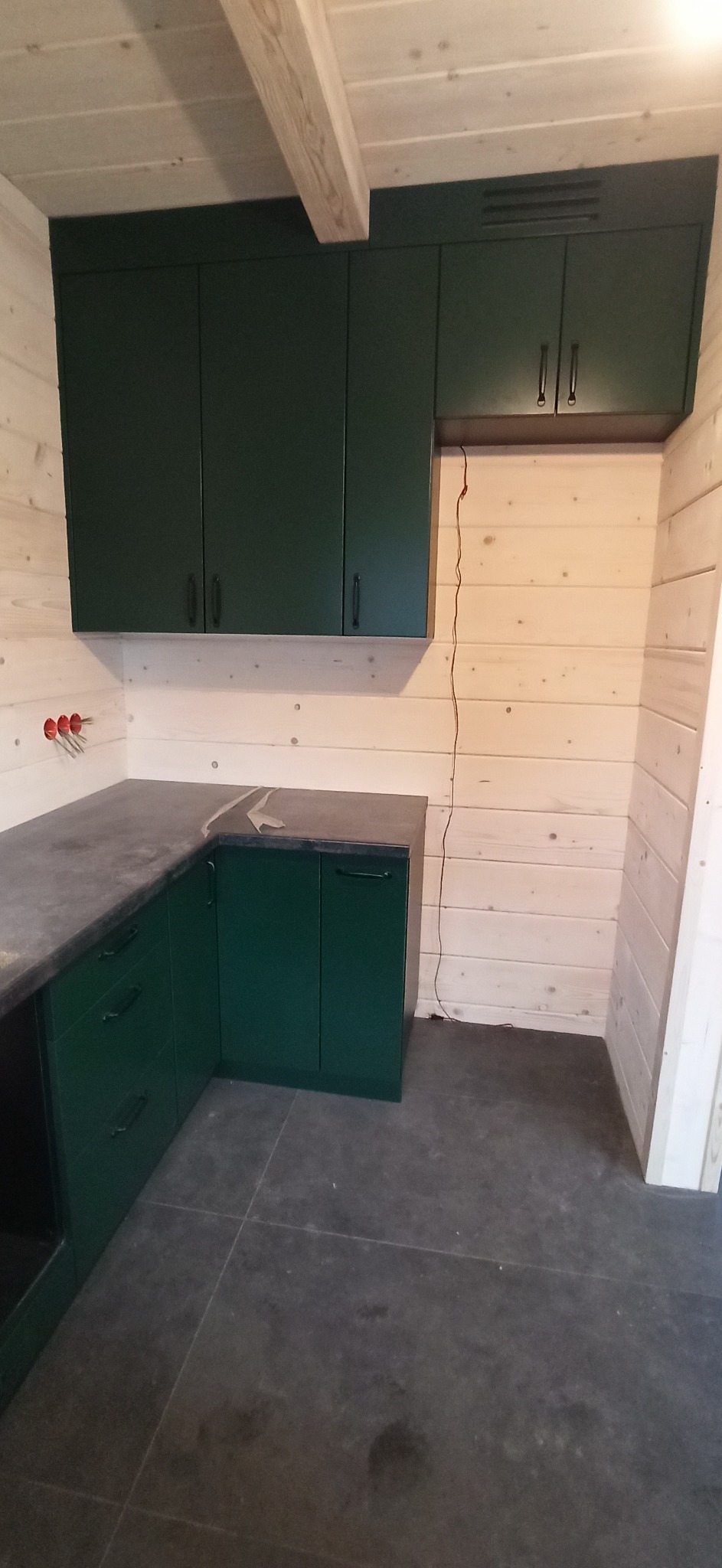 Kącik kuchenny z ciemnozielonymi szafkami, szarym blatem i drewnianą boazerią ścienną w surowym stylu.