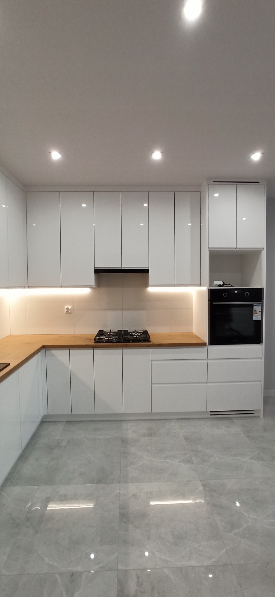 Biała zabudowa kuchenna z drewnianym blatem, oświetleniem LED pod szafkami wiszącymi, płyta gazowa i piekarnik w zabudowie, szara marmurkowa podłoga.