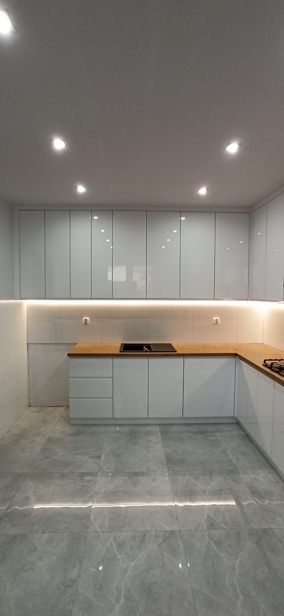 Nowoczesna, jasna kuchnia z białymi, lakierowanymi szafkami, drewnianym blatem i szarą podłogą z płytek imitujących marmur, oświetlenie LED podszafkowe.