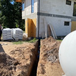 Instalacja24 Damian Sikora - Wykop pod rury przyłącza do budynku w stanie surowym, widoczne niebieskie rury w ziemi, worki z zaprawą murarską i częściowo zbiornik na szambo.