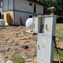 Instalacja24 Damian Sikora - Dwuczęściowa, otwarta skrzynka elektryczna TN-C z ostrzeżeniem o napięciu, stojąca na piaszczystym gruncie budowlanym przed domem w stanie surowym, z widocznym zbiornikiem gazu, w tle drzewa...