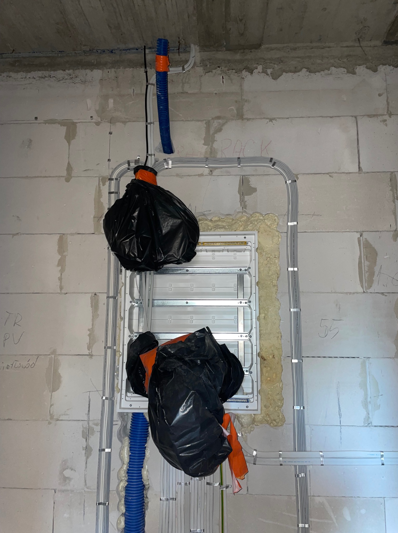 Rozdzielnica elektryczna w trakcie montażu, zabezpieczona czarnymi workami foliowymi, widoczne kable w peszlach i rurkach mocowane do ściany z betonu komórkowego.