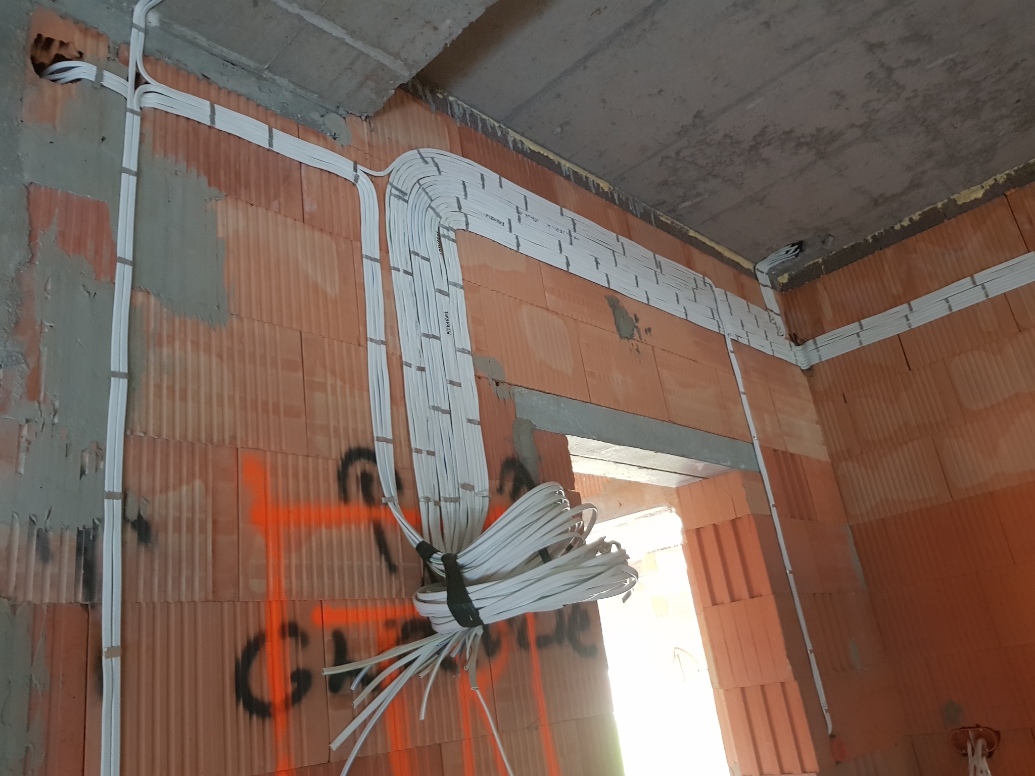 Wiązka białych kabli elektrycznych prowadzonych w peszlach, przymocowanych do ściany z czerwonej cegły dziurawki w surowym stanie, z widocznymi oznaczeniami i śladami po pracach budowlanych.