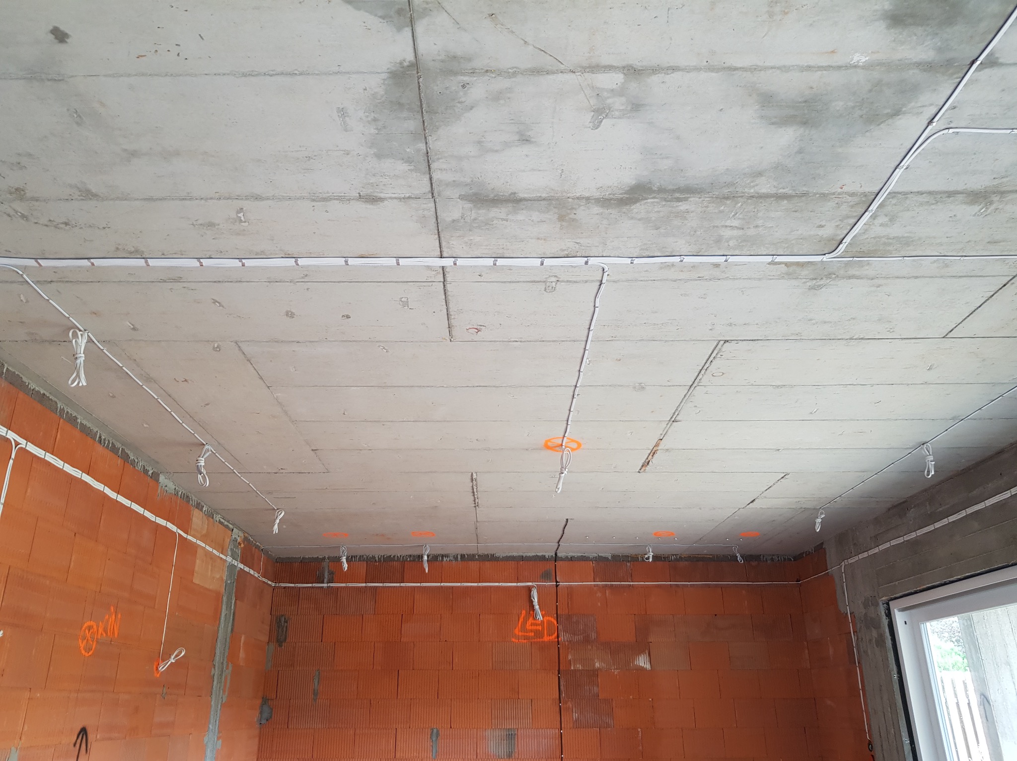 Surowy betonowy strop z widocznymi przewodami elektrycznymi, częściowo zamocowanymi do ściany z czerwonej cegły, z pomarańczowymi oznaczeniami. Przewody zwisają na sznurkach.