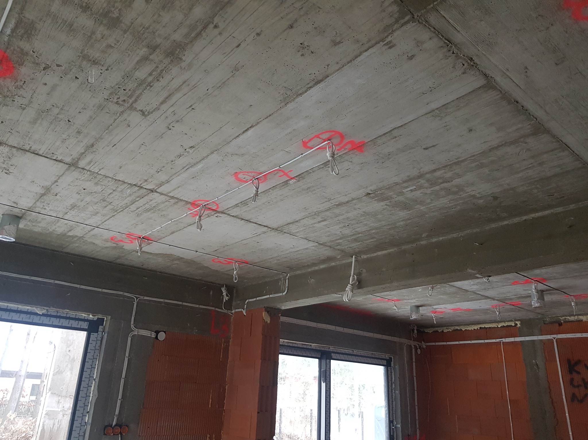 Surowy betonowy strop z widocznymi przewodami elektrycznymi, oznaczonymi czerwonymi numerami i zawieszkami, w trakcie prac instalacyjnych w nowym budynku.