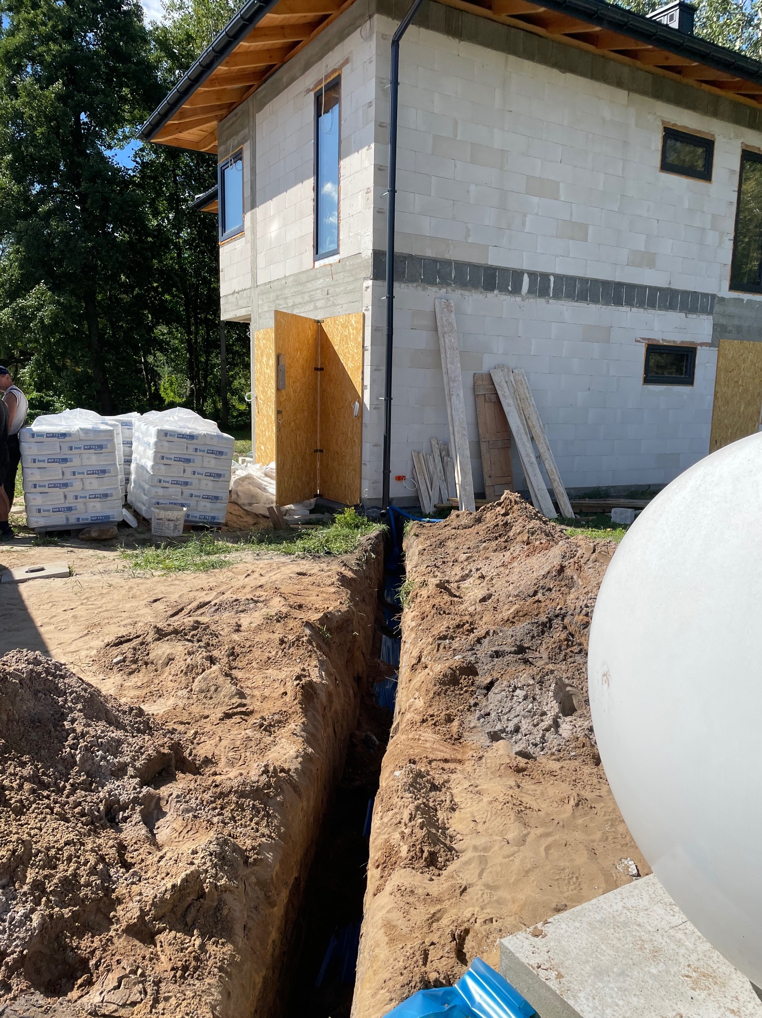 Wykop pod rury przyłącza do budynku w stanie surowym, widoczne niebieskie rury w ziemi, worki z zaprawą murarską i częściowo zbiornik na szambo.